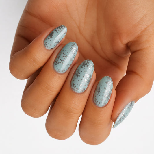 Pale sage-blue nail wraps with silver and white snowflake patterns, glossy finish, almond shape