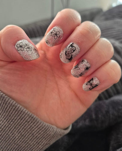 Hand wearing silver glitter gel nail wraps with black butterfly nail art patterns